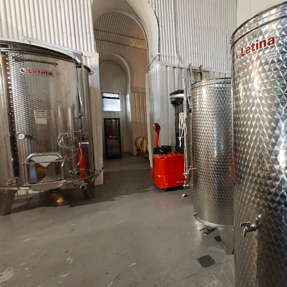 Large stainless steel wine fermenters and tanks inside a winery with concrete floors, corrugated metal walls, and an orange cart in the background. The brand name 'Letina' is visible on the tanks.
