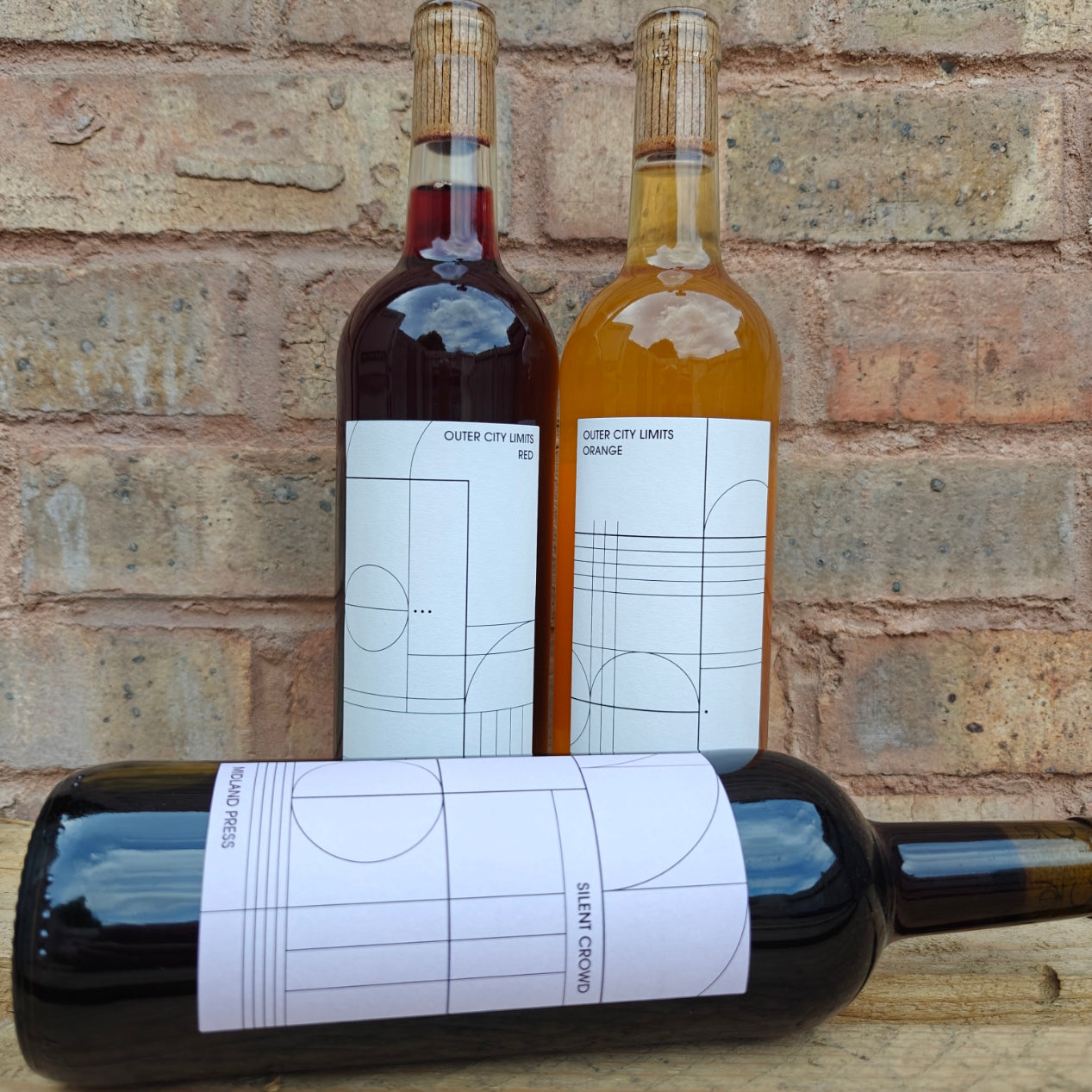 Three wine bottles with minimalist white labels featuring geometric line art, set against a rustic brick wall background. One bottle is lying horizontally in the foreground, while two stand upright behind it.