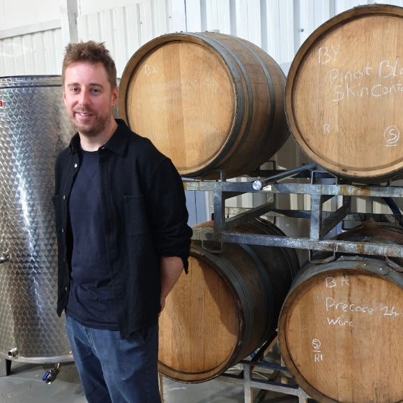 Richard Stenton, the winemaker, standing in a winery next to multiple wooden barrels on metal racks, with a stainless steel vat visible on the left.